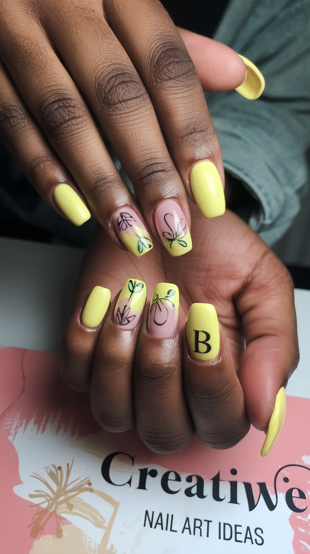 Image of hands with butter yellow nails featuring floral designs and a letter, placed on a creative nail art ideas background.
