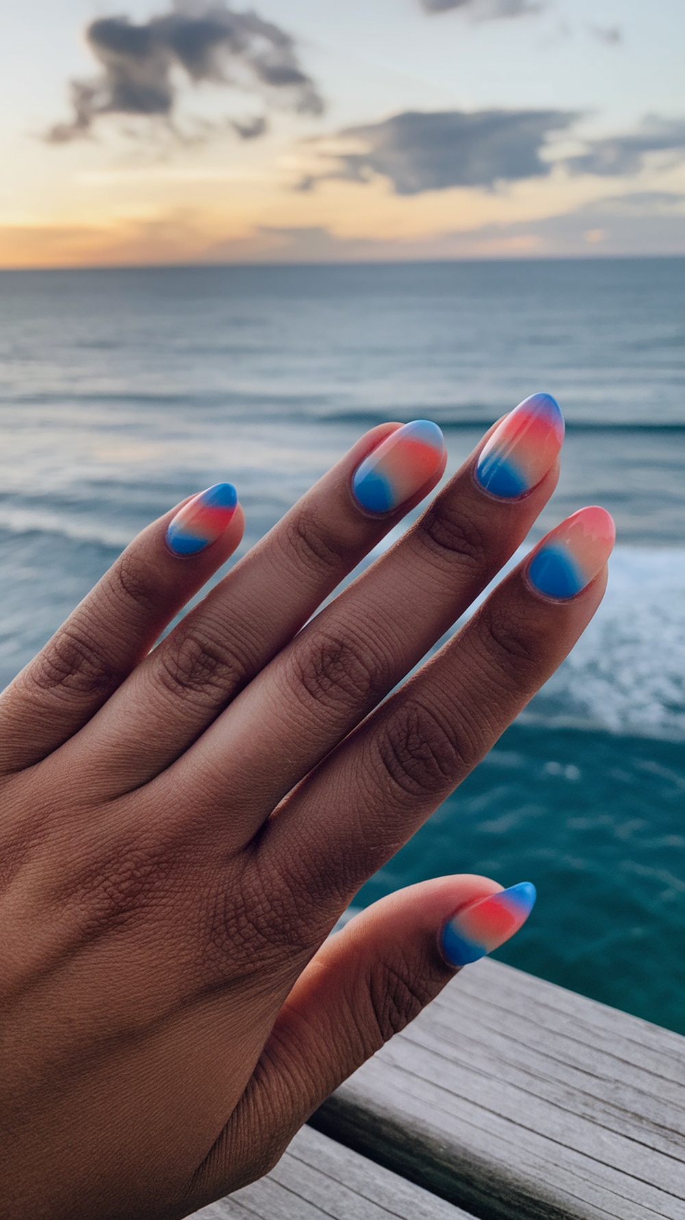 A close-up of beach sunset gradient nails with pink and blue hues against a seaside background.