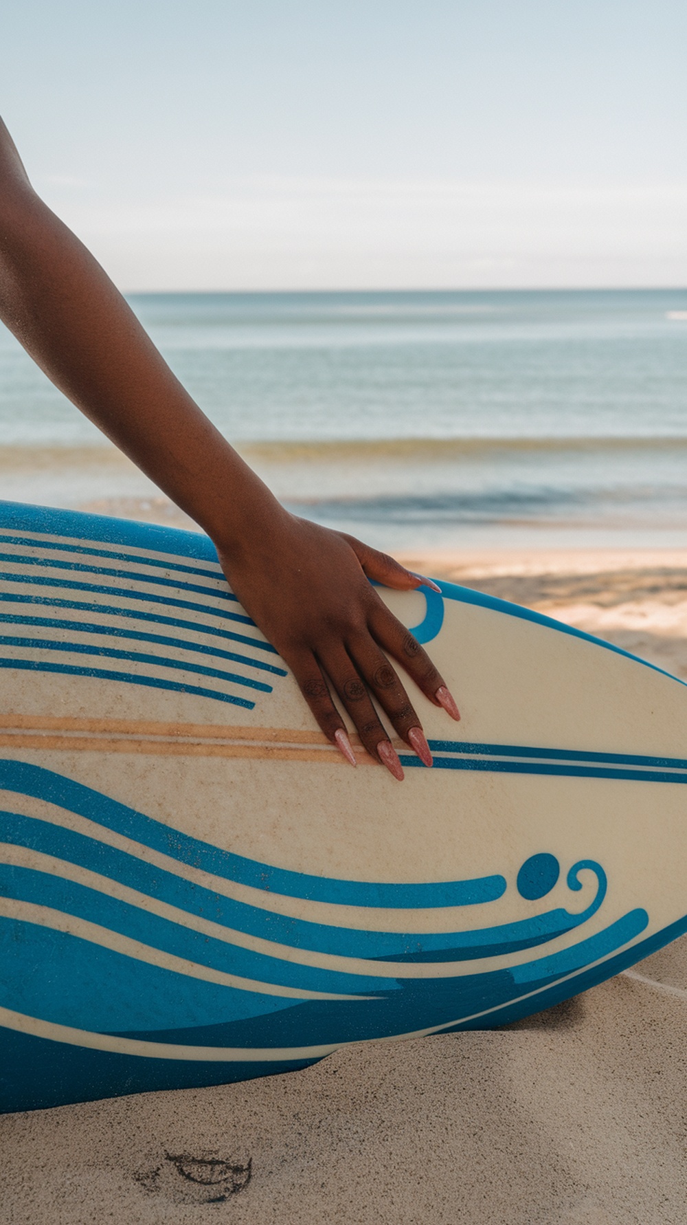 A hand resting on a surfboard on the beach, showcasing summer vacation nails.