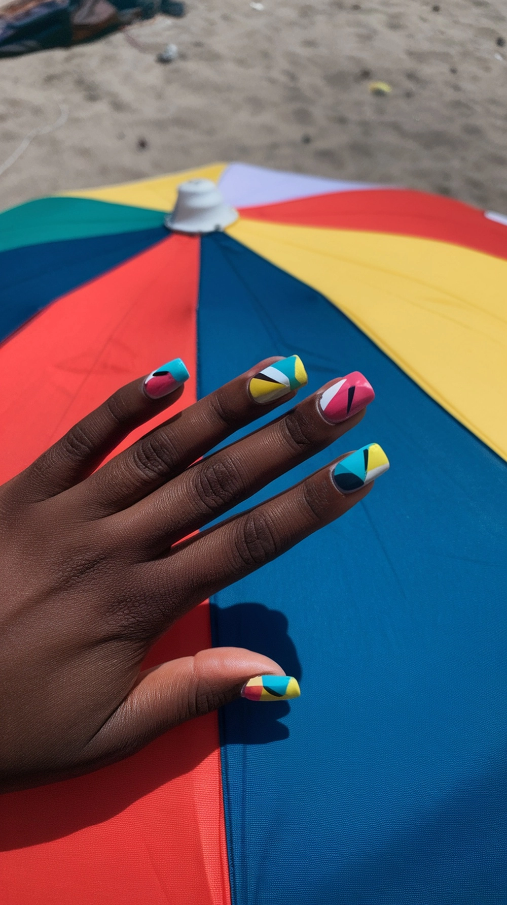 Colorful geometric nail designs on a hand with a beach umbrella in the background.