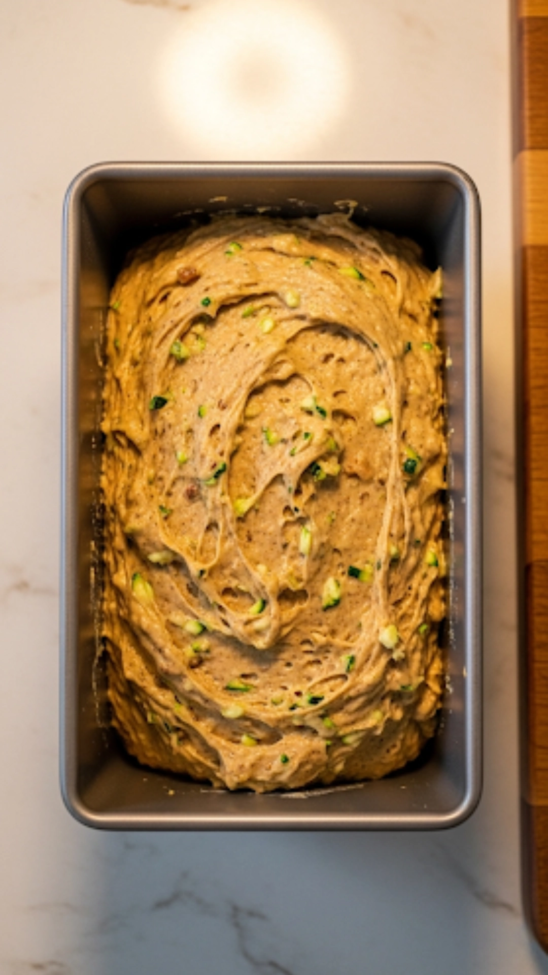 Thick zucchini bread batter with visible flecks of green zucchini, filled in a metal loaf pan and ready to be baked.