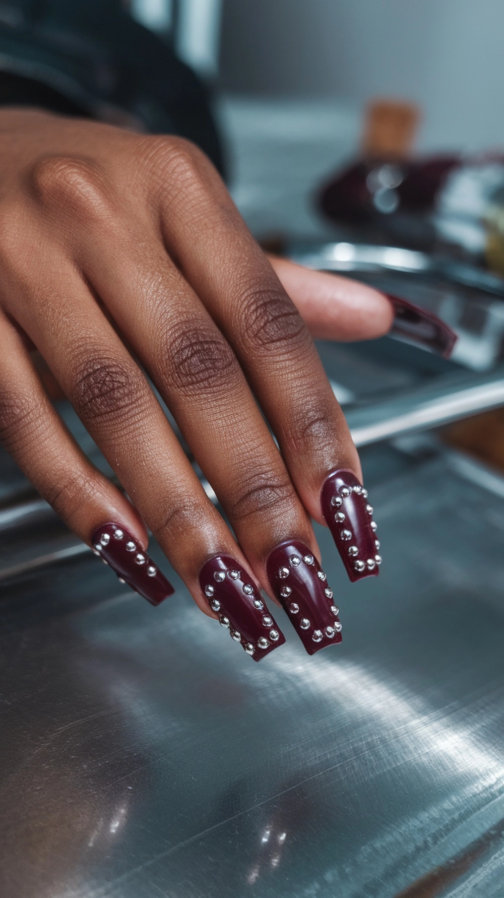 How to Style Burgundy Nail Designs for a Bold Stunning Look 3 Burgundy nails with silver studs on a hand resting on a metallic surface.