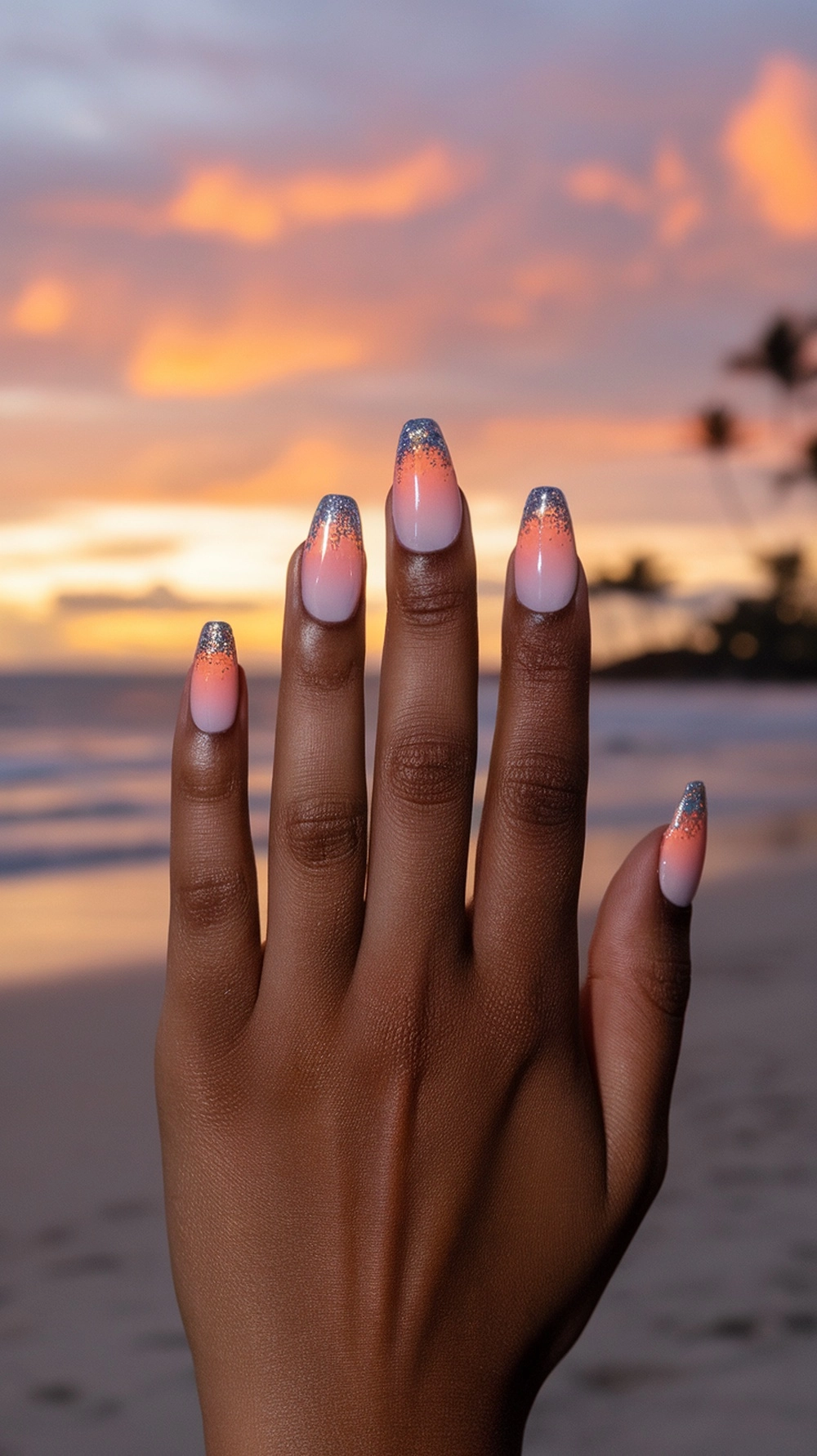 16 Cute Boho Nail Ideas You’ll Love 10 A hand with sunset ombre nails featuring glitter tips, set against a beach sunset.