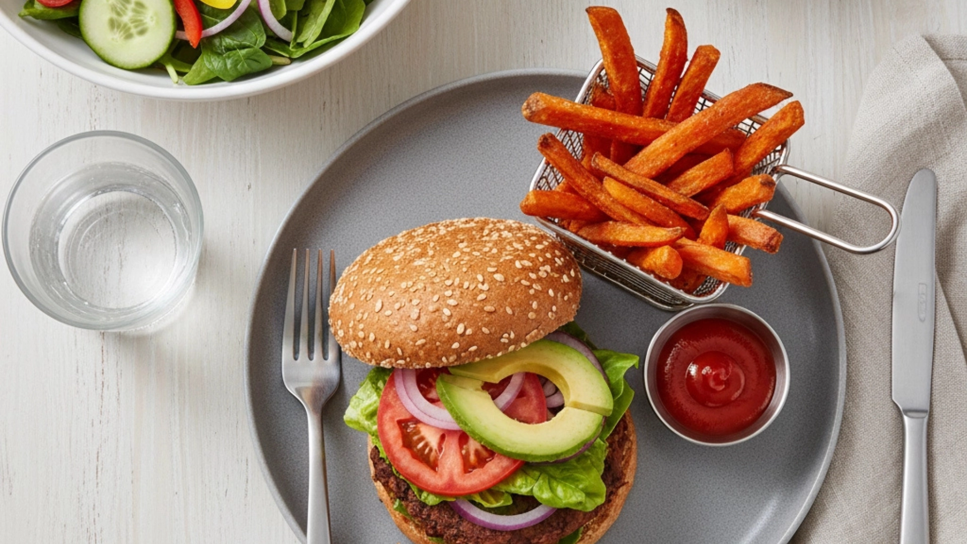A vibrant flat lay of a healthy weeknight meal on a white wooden table. A plated black bean burger with avocado and tomato sits centrally, flanked by a bowl of crispy sweet potato fries and a fresh green salad.