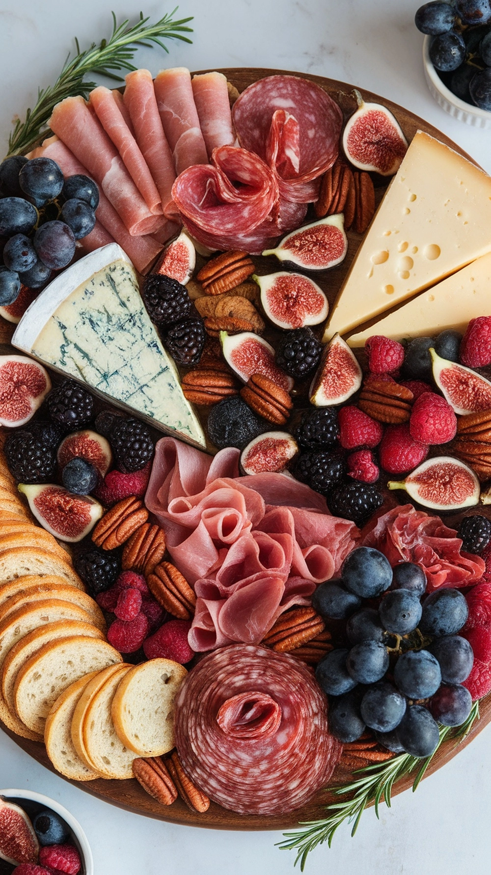 A top-down view of a round wooden charcuterie board filled with cured meats, blue cheese, grapes, figs, pecans, and sprigs of rosemary.