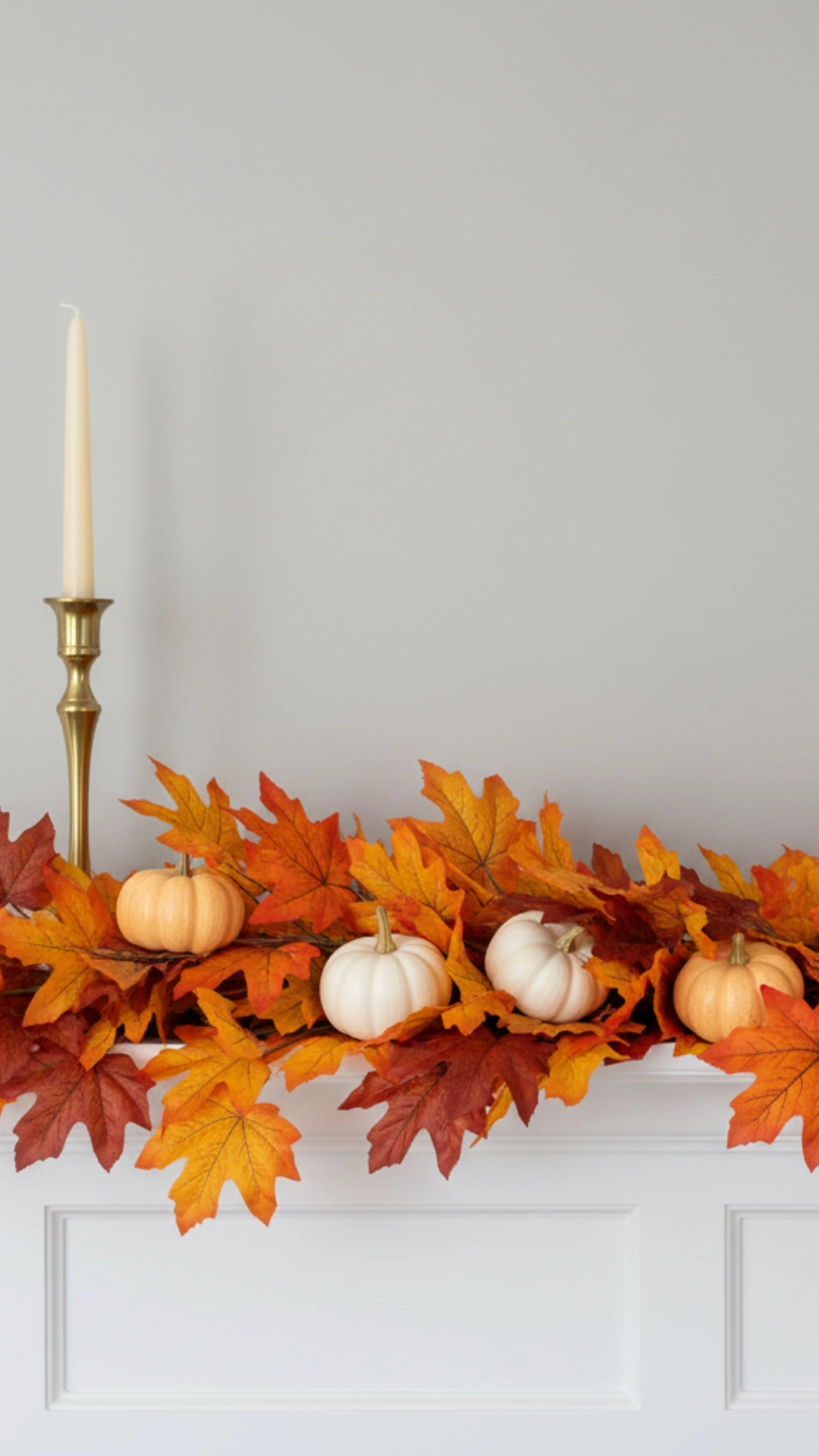 How to Use Cozy Home Decor to Unleash the Power of Fall 7 A close-up of a beautifully decorated white fireplace mantle. It features a garland of autumn leaves, tall brass candlesticks with cream candles, and an assortment of small white and orange pumpkins nestled within the foliage.