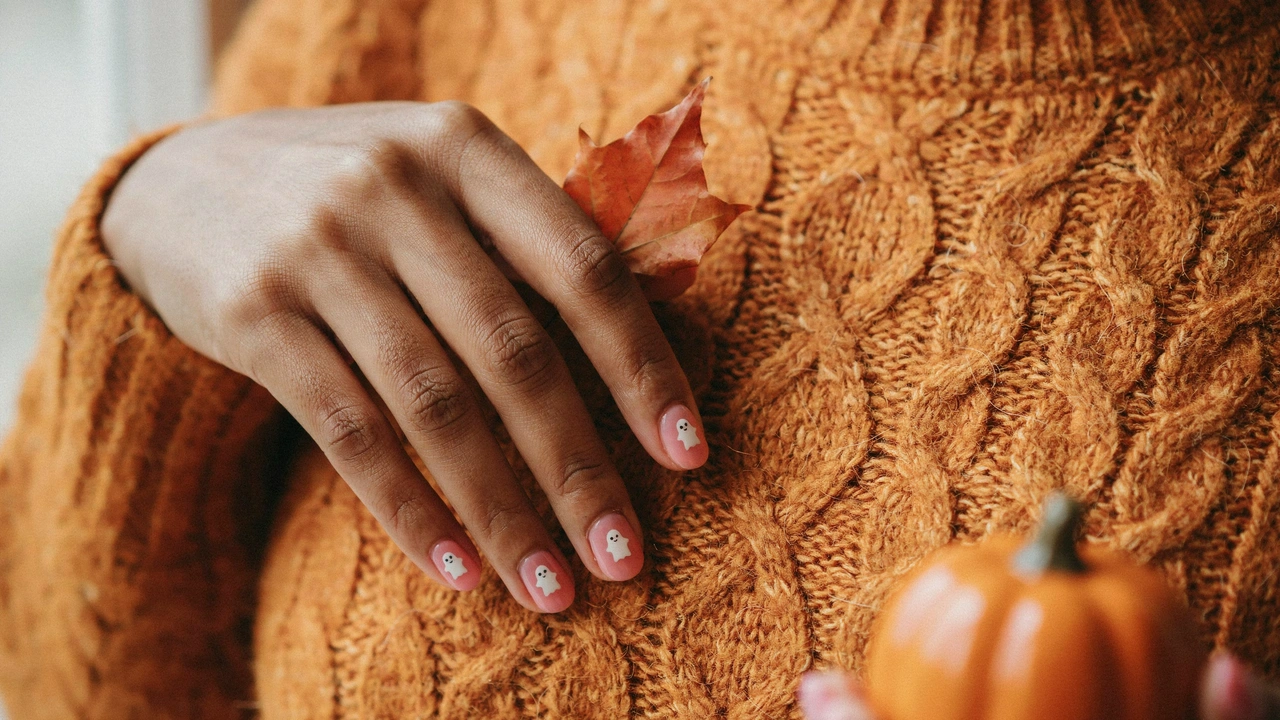 A dark brown woman's hand resting on an orange sweater showing off simple Halloween nail art featuring tiny white ghosts.