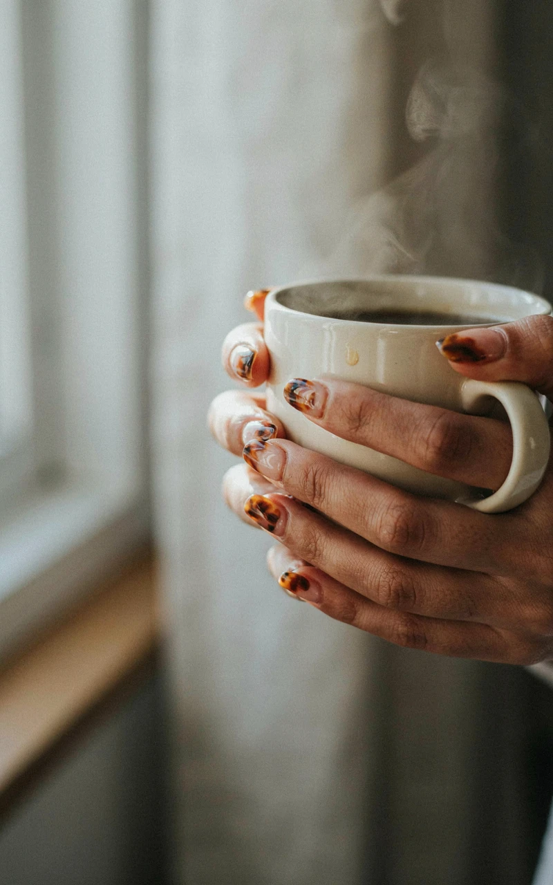 13 Almond Fall Nail Ideas That Look Elegant and Trendy 2 A dark brown woman's hand holding a mug with tortoiseshell painted on the tips of her almond nails.
