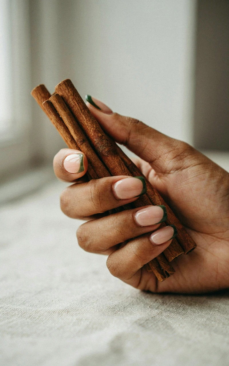 13 Almond Fall Nail Ideas That Look Elegant and Trendy 6 A dark brown woman's hand holding cinnamon sticks with forest green French tips on her almond nails.