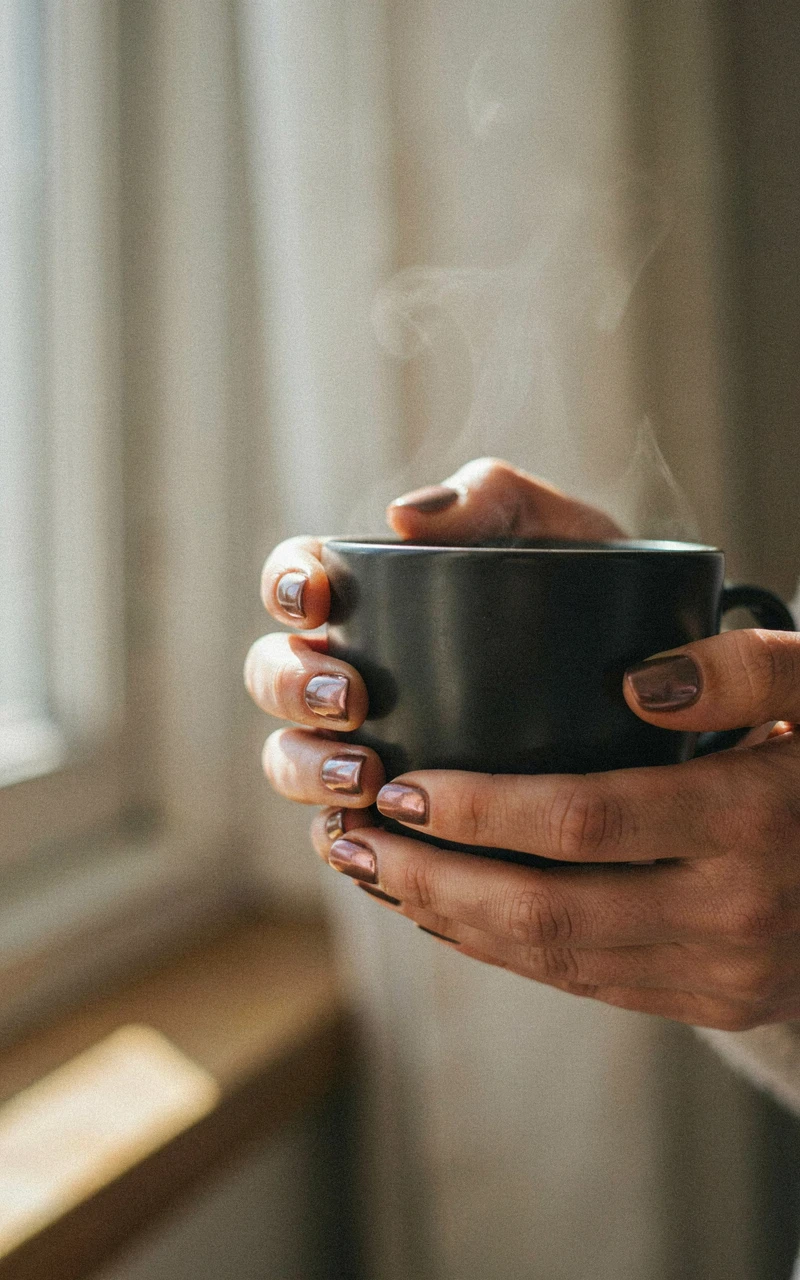 13 Almond Fall Nail Ideas That Look Elegant and Trendy 10 A dark brown woman's hand holding a coffee cup with pearlescent chocolate brown almond nails.