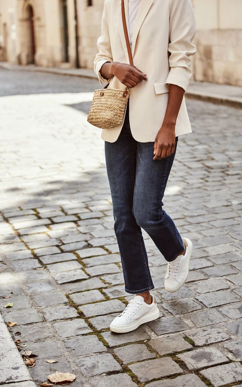 A close-up of a dark brown woman wearing a cream blazer, adjusting her crossbody bag while wearing white sneakers.