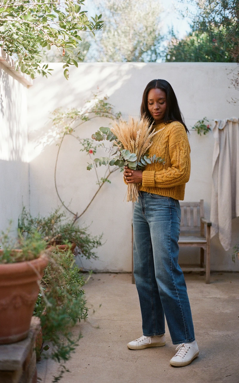 A dark brown woman wearing a mustard yellow cropped sweater and high-waisted jeans holding dried flowers.