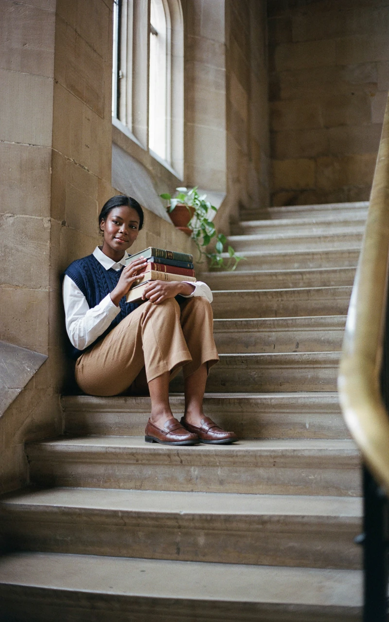 A dark brown woman wearing a navy cable knit vest over a white button-down shirt holding books.