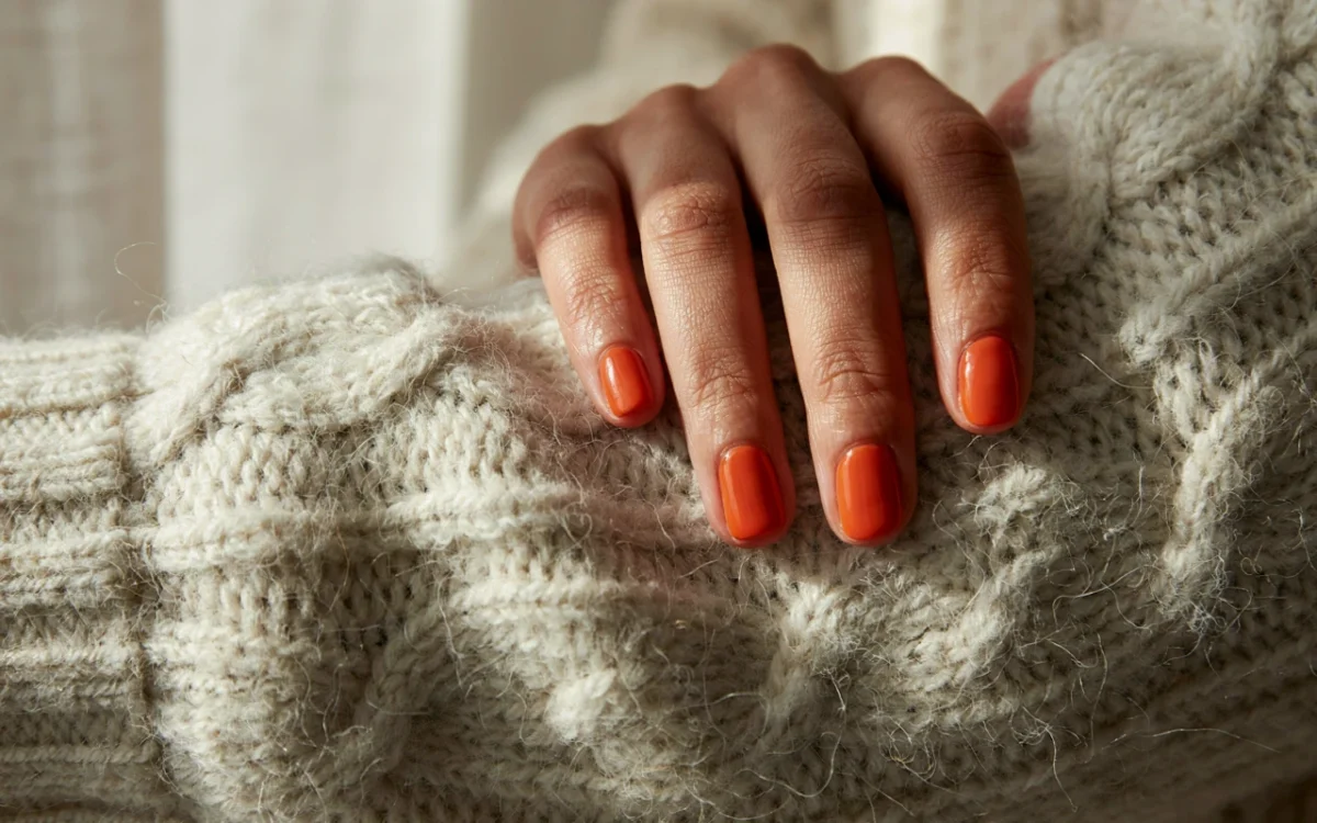 A dark brown woman's hand resting on a knit sweater showing off glossy burnt orange short fall nail ideas.