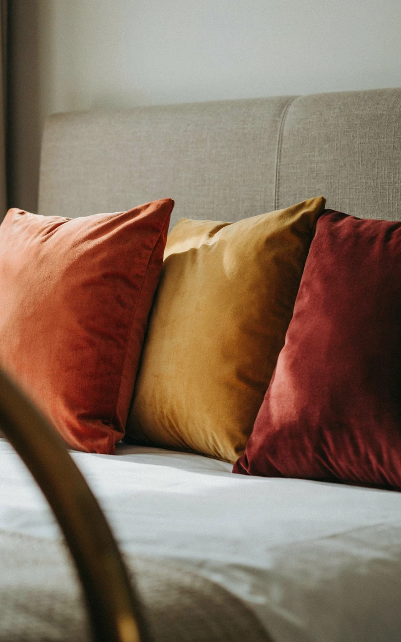 Three velvet throw pillows in rust, mustard, and burgundy on a bed.
