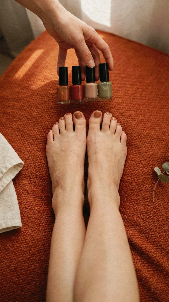 A woman's hand holding brown, burgundy, taupe and olive polish bottles above the feet with brown toe nails on a rust rug