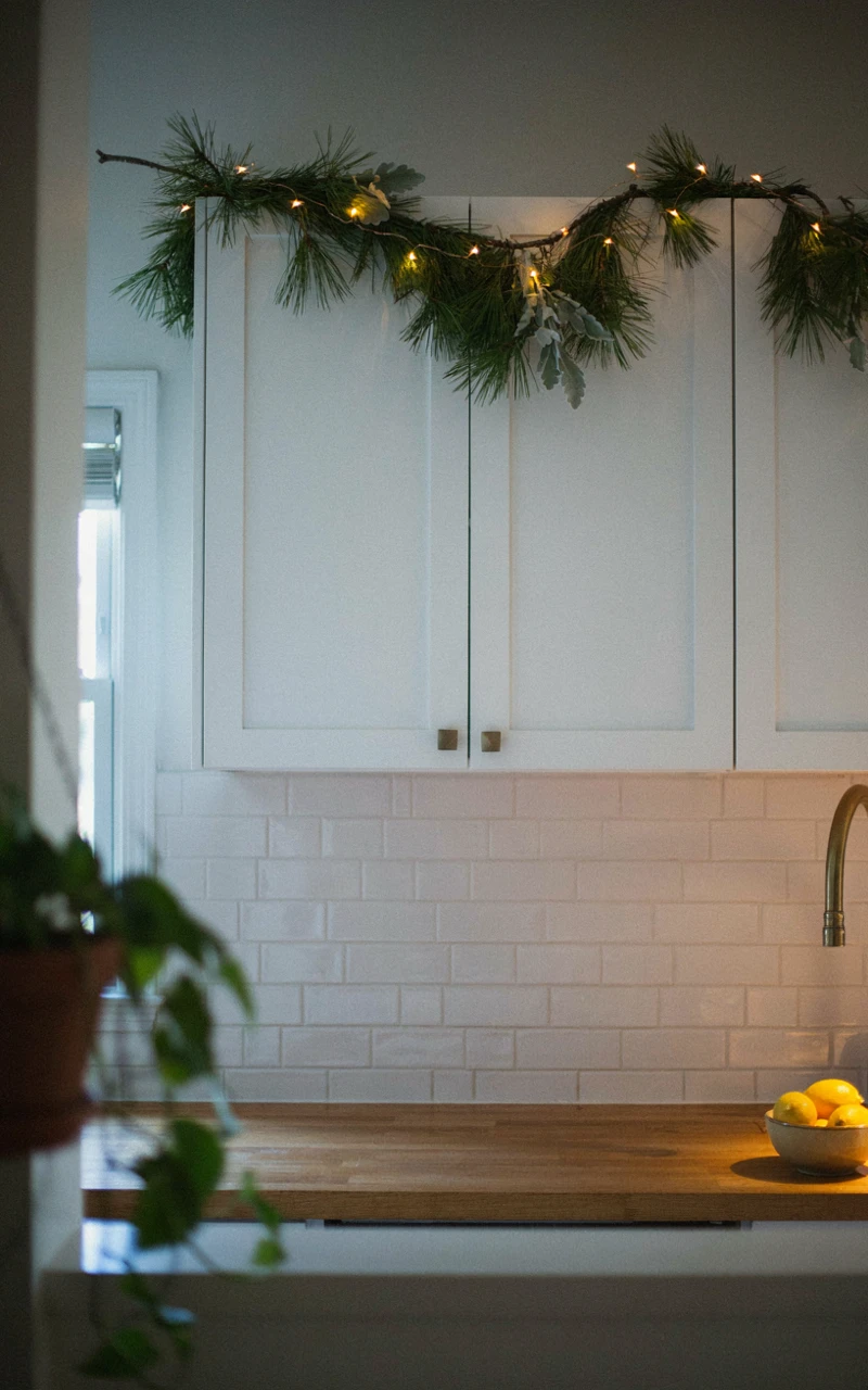 13 Christmas Kitchen Decor Ideas for a Warm and Festive Home 4 Warm string lights and a pine garland glowing along the tops of white kitchen cabinets in the evening