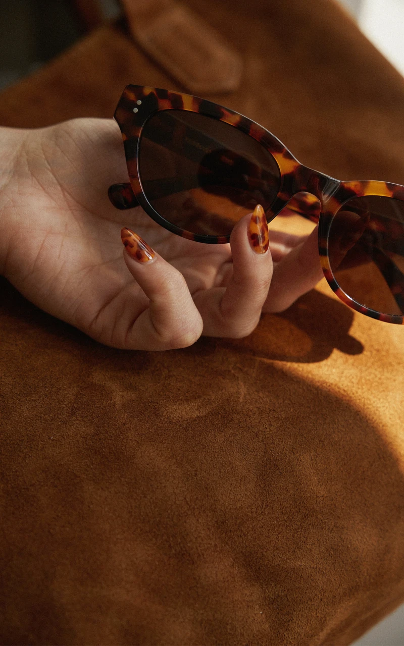 Tortoiseshell brown chrome nails on a woman's hand holding tortoiseshell sunglasses against a brown suede bag