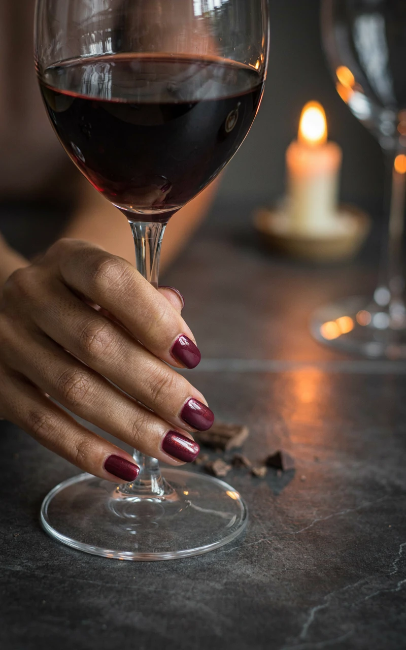 Burgundy chrome nails with a ruby glint on a woman's hand holding a red wine glass by candlelight