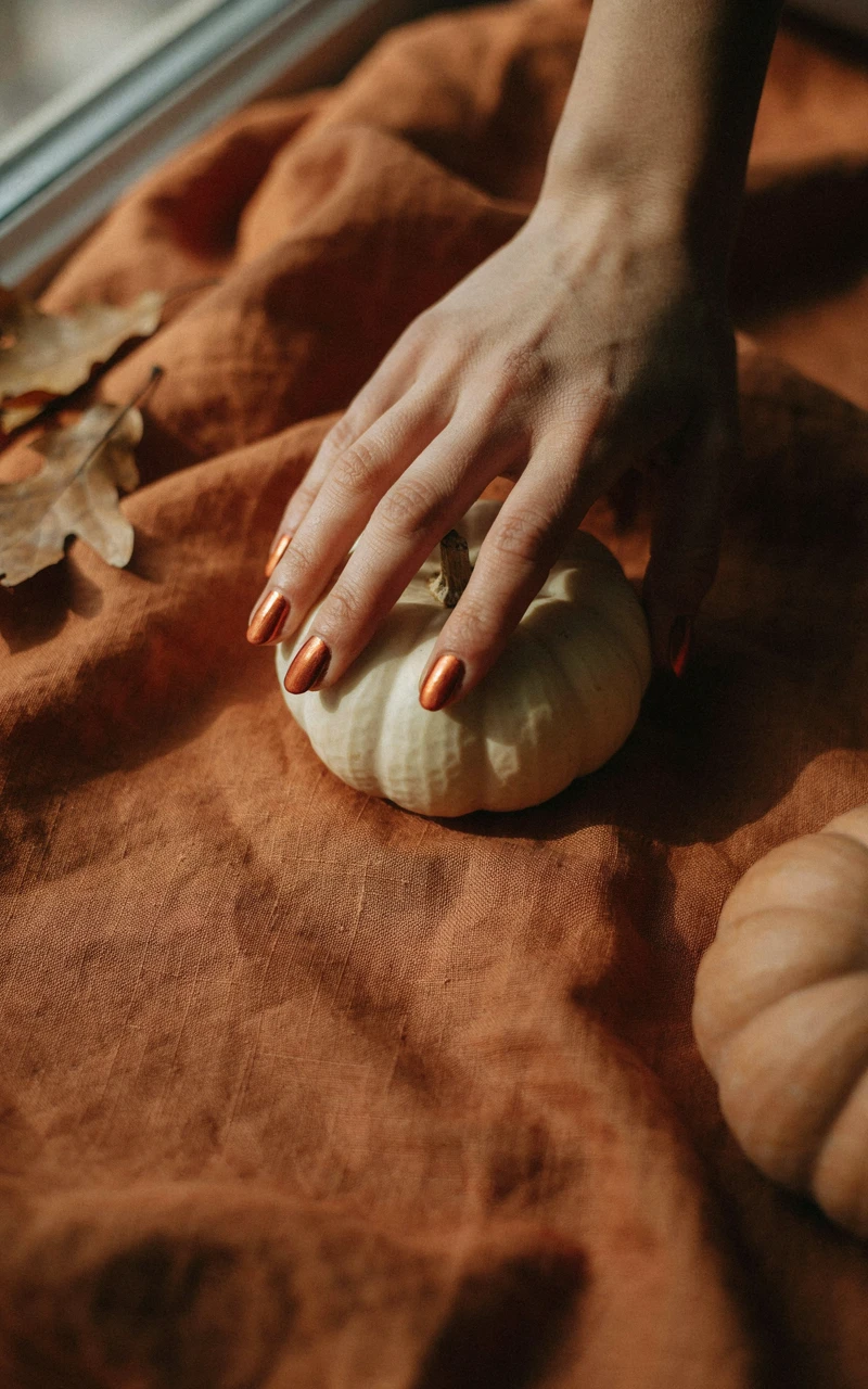 Burnt orange autumn chrome nails on a woman's hand resting on a small white pumpkin on a rust linen throw