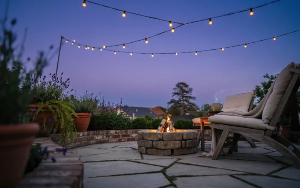 A dusk patio full of cozy backyard ideas: string lights, a stone fire pit, and cushioned chairs with a knit throw