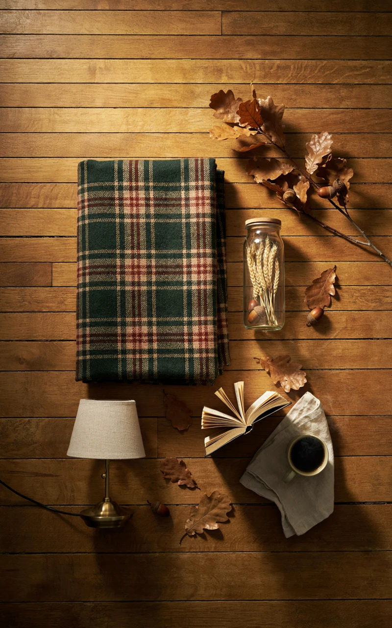 How to Create a Cozy Autumn Bedroom Without Buying Lots of Decor 2 A folded plaid blanket, a glass jar, dried branches, and a small brass lamp on a wooden floor.