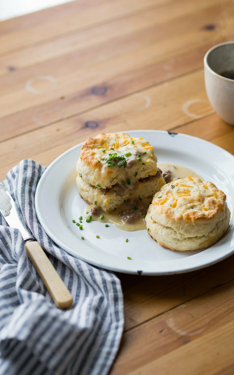 10 Cozy Fall Breakfast Ideas for Weekend Mornings 7 A split cheddar chive biscuit covered in creamy sausage gravy and chives on a white plate for a cozy breakfast