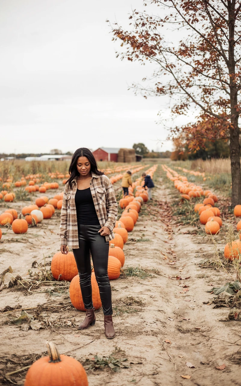 12 Cozy Fall Outfits for Women That Feel Stylish and Comfortable 5 A woman in a pumpkin patch wearing a plaid shacket and black faux leather leggings.