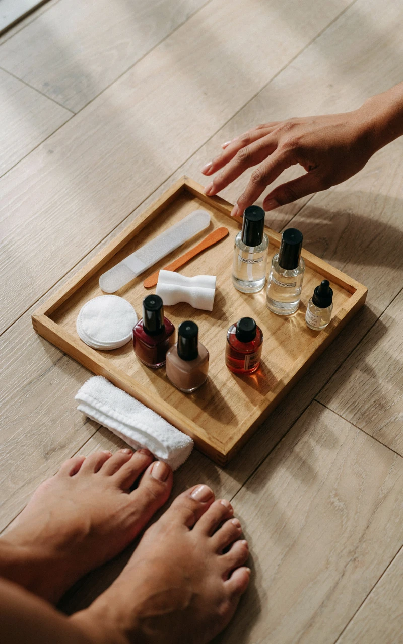 A woman's hand reaching into a wooden tray of pedicure tools, toe separators, base coat and brown, burgundy and taupe polish