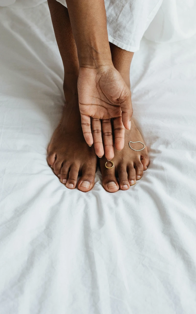 A woman's wrist and bare unpainted toenails in daylight beside a gold and a silver bracelet on a white linen sheet