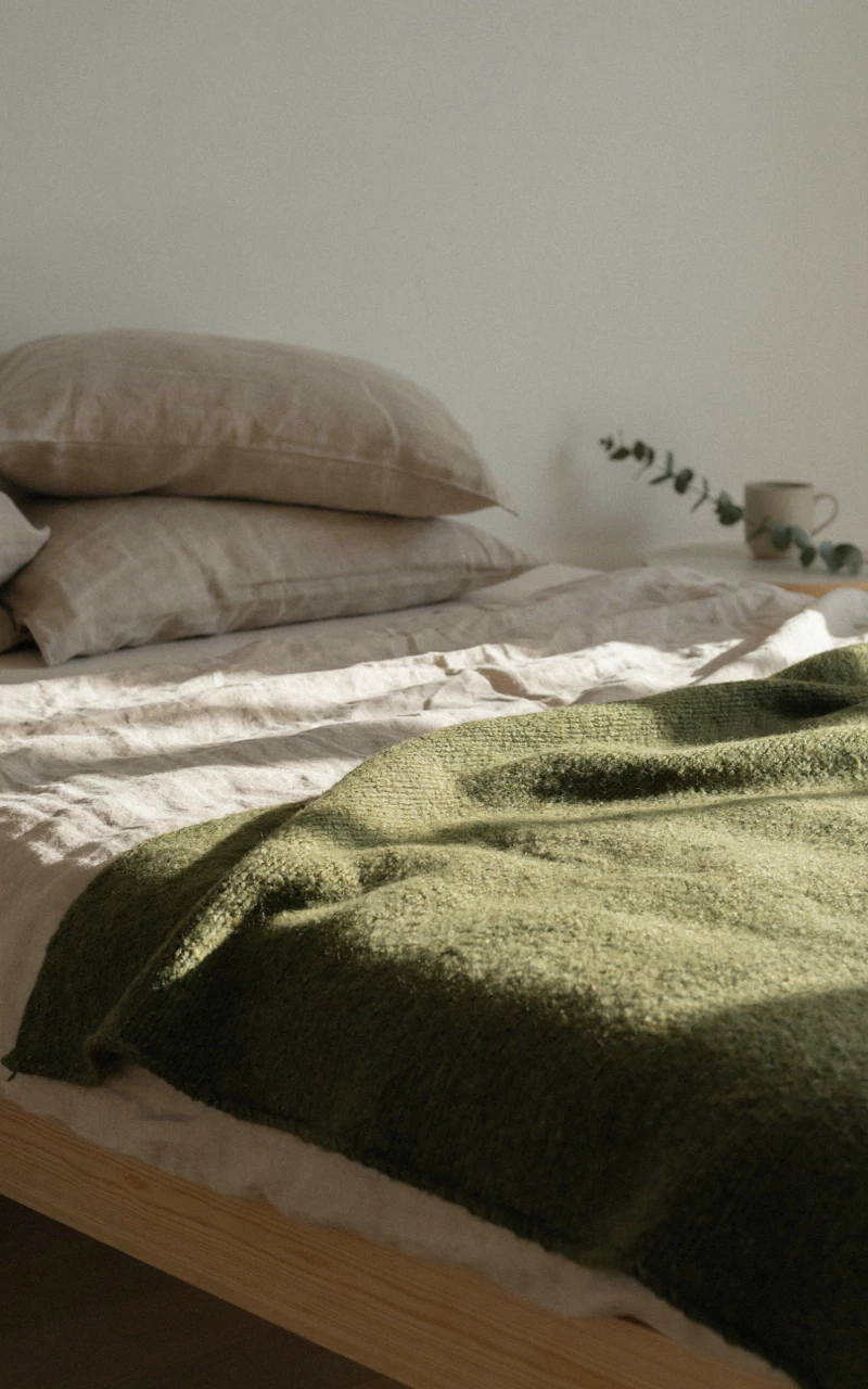 17 Green Aesthetic Bedroom Ideas That Feel Cozy and Stylish 16 Chunky moss green knit blanket draped across the foot of a bed with oatmeal linen bedding in afternoon light