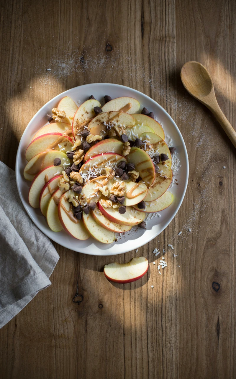 11 Healthy Snack Ideas for Any Time of Day 8 Apple nachos drizzled with almond butter and topped with chocolate chips, walnuts and coconut on a white plate