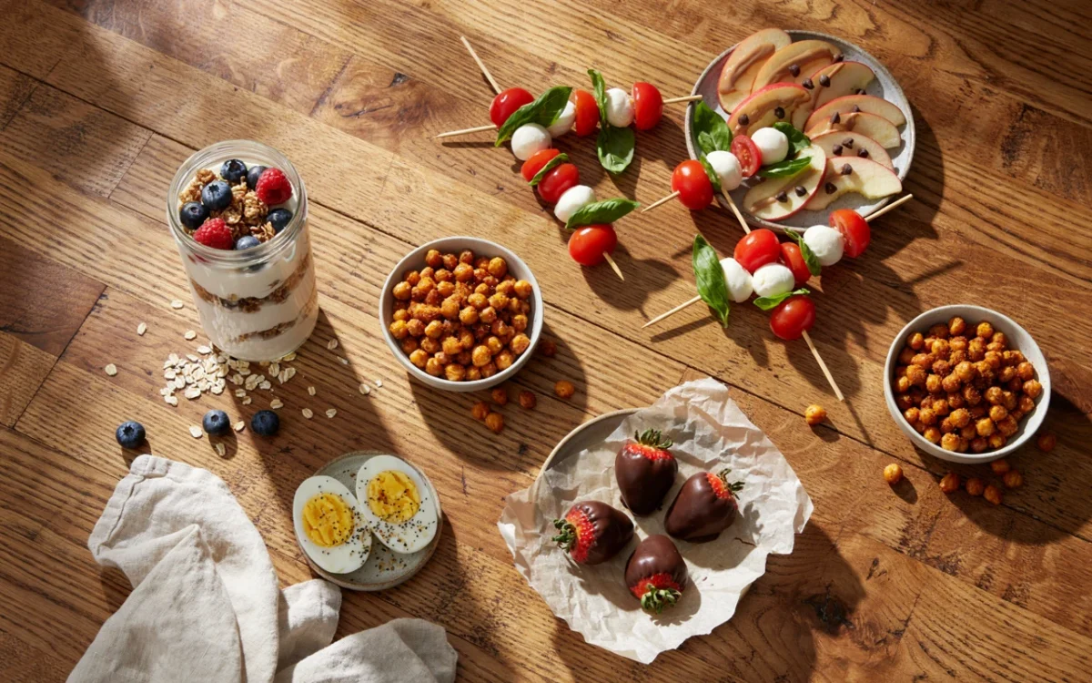 A wooden table of healthy snack ideas: yogurt parfait, roasted chickpeas, apple slices, caprese skewers and eggs