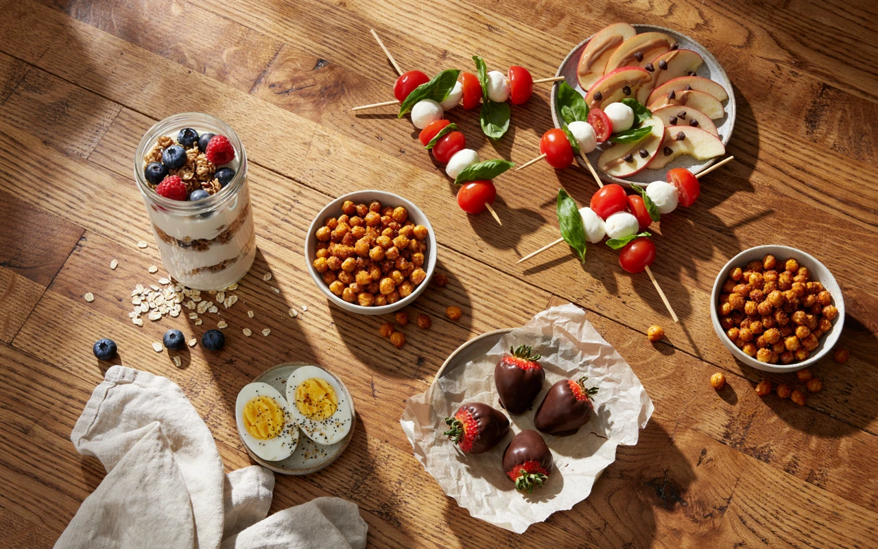 11 Healthy Snack Ideas for Any Time of Day 1 A wooden table of healthy snack ideas: yogurt parfait, roasted chickpeas, apple slices, caprese skewers and eggs