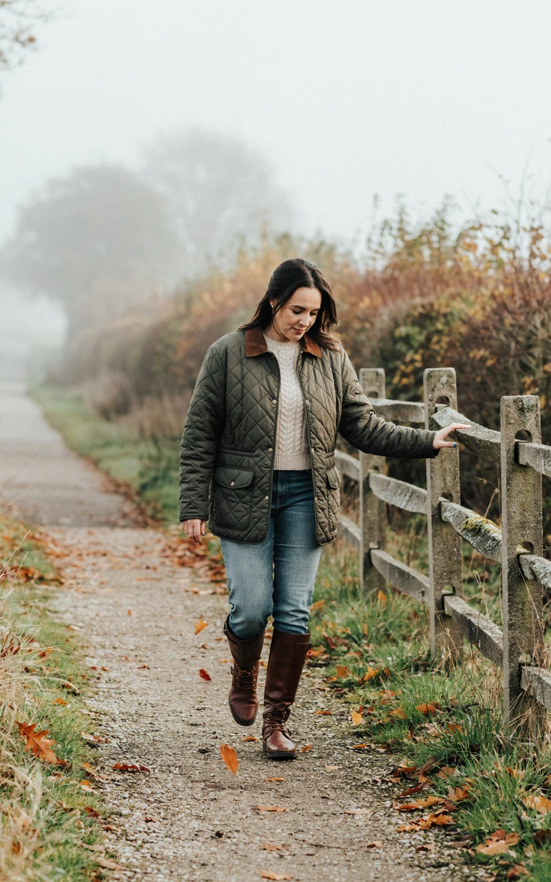 A Woman in an olive quilted jacket, straight jeans and tall brown riding boots on a fall country lane