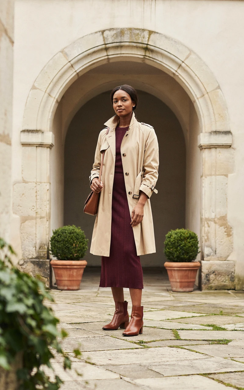 A Woman in a belted beige trench coat over a burgundy knit midi dress with brown ankle boots