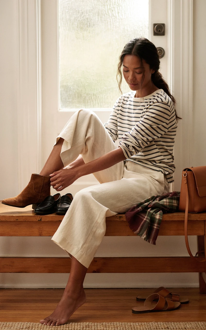 A woman puts on suede ankle boots on an entryway bench, with loafers, a plaid scarf and a leather bag beside her